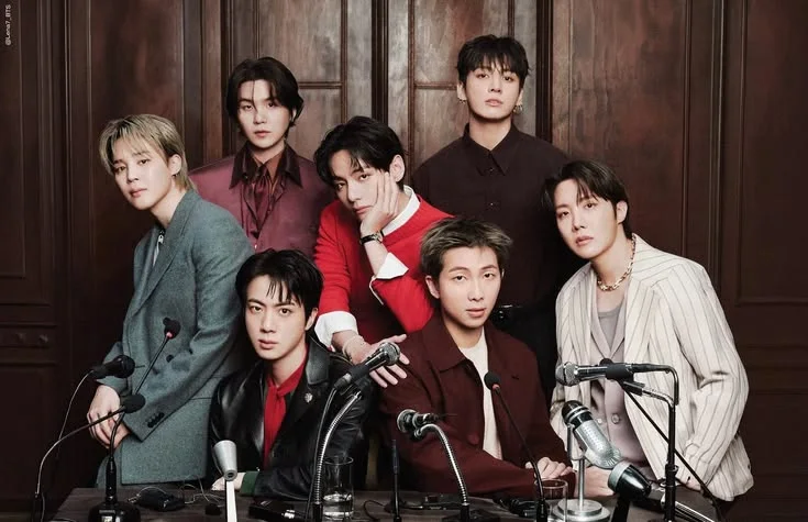 Os sete integrantes do BTS reunidos atrás de uma mesa com microfones